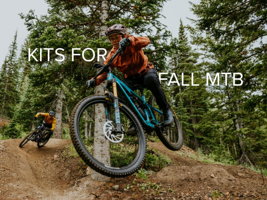 Two MTB riders navigate a jump and a berm, respectively, during a soggy fall ride in an alpine forest. 